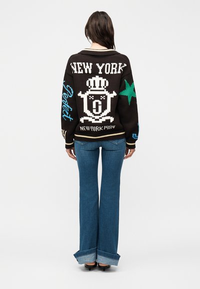 Femme debout de dos, portant un pull noir en tricot avec l'inscription « NEW YORK », un emblème couronné et du texte, associé à un jean bleu retroussé et des talons noirs.