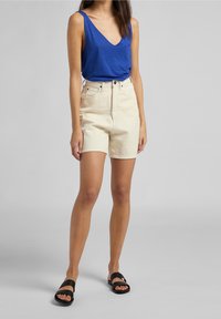 Top sin mangas azul, pantalones cortos beige de talle alto y sandalias negras. Los pantalones cortos tienen un corte recto y una textura suave.