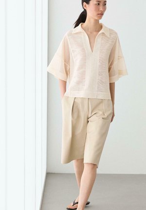 Frau steht am Fenster, trägt ein durchsichtiges cremefarbenes, strukturiertes Oberteil, beige knielange Shorts und schwarze Sandalen, blickt nach rechts mit den Händen in den Taschen.