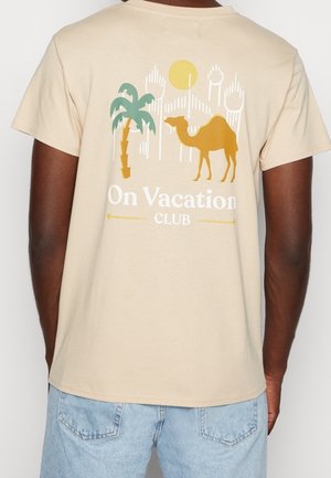 T-shirt beige avec un design graphique représentant un chameau, un palmier et un soleil. Le texte indique "On Vacation CLUB" en blanc. Manches courtes, col rond.