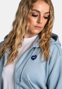 Donna con lunghi capelli biondi ondulati che guarda in basso, indossa una camicia bianca e una felpa leggera con zip di colore azzurro chiaro con un logo a forma di cuore di Carhartt WIP.
