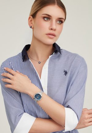 Donna con trucco leggero indossa una camicia a righe blu e bianche, colletto blu scuro, collana e orecchini floreali e un orologio da polso in argento con quadrante blu.