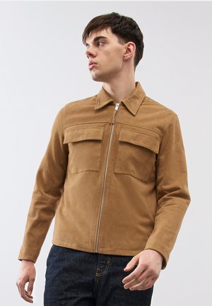 Giovane uomo rivolto verso sinistra che indossa una giacca marrone chiaro con cerniera e tasche con patta e jeans blu scuro, su uno sfondo chiaro e semplice.