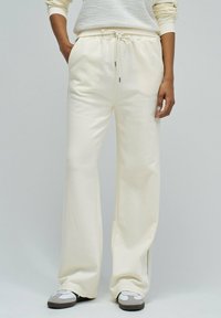 Personne portant un pantalon de survêtement large crème avec une taille coulissante, associé à des baskets blanches et grises, se tenant devant un fond uni.