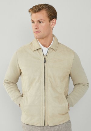 Beige jacka med en texturerad, mockaliknande ovansida, stickade ärmar, framdragkedja och sidofickor. Minimalistisk design med horisontell sömnad.