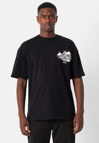 Camiseta de algodón negra con un fit relajado, que presenta un diseño gráfico blanco en el lado izquierdo del pecho, manga corta y cuello redondo.