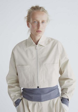 Mujer rubia con ojos azules que lleva una chaqueta corta beige sobre una camiseta gris, de pie con las manos en los bolsillos frente a un fondo liso.