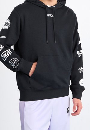 Personne portant un sweat à capuche noir avec des imprimés graphiques blancs sur les manches et la poitrine, associé à un pantalon violet clair avec une petite étiquette logo noire.