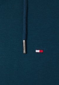 Sudadera color teal con una tela texturizada, que presenta una cremallera de metal plateado con la inscripción "HILFIGER" y un pequeño logo bordado en rojo, blanco y azul.