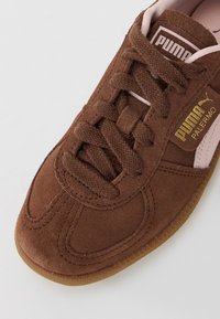 Brun mocka Puma Palermo sneakers med bruna snören, rosa detaljer och guld "Puma Palermo"-logotyp på vit bakgrund.