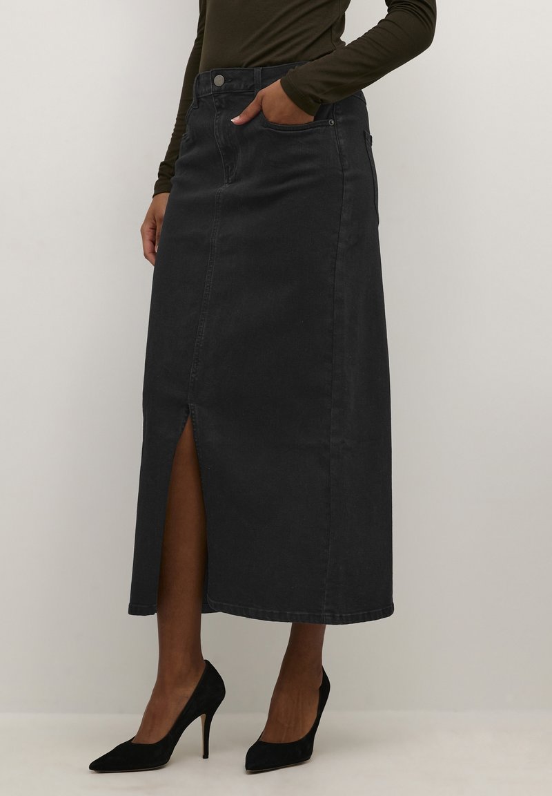 Femme portant une jupe midi en jean noir avec une fente à l'avant, des talons hauts noirs et un haut sombre à manches longues, debout devant un fond uni.