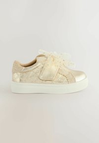 Goldfarbene metallische Kindersneaker mit gesticktem Muster, Glitzerelementen und großer dekorativer Schleife auf weißer Sohle vor weißem Hintergrund.