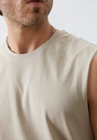 Light beige sleeveless shirt, cotton material, round neckline, smooth texture, subtle stitching detail, worn with a silver chain necklace.