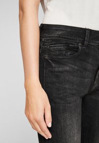 Jean en denim noir avec une texture décolorée, un design à cinq poches et des coutures visibles. Gros plan sur la taille avec une main posée dessus.