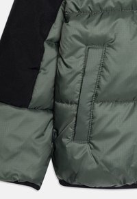 Grön puffjacka med en texturerad yta, med en ärm av svart ull-liknande tyg och en dragkedjeficka på bröstet.