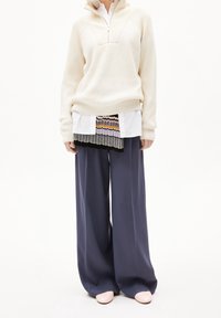 Hoodie crème côtelé à demi-fermeture éclair porté sur une chemise blanche, superposé avec un haut à motif et un pantalon large bleu marine, assorti à des chaussures roses.