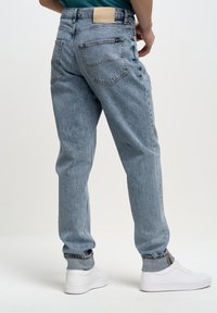 Jeans de mezclilla azul claro con un corte relajado, diseño de cinco bolsillos y costuras a contraste. La cinturilla presenta un parche de cuero color beige claro.