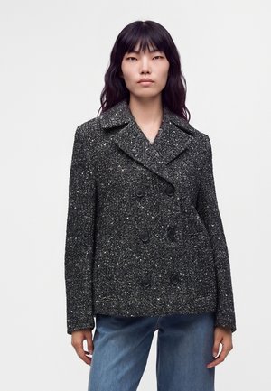 Kvinde iført en mørkegrå dobbeltradet frakke med fiskebenmønster og sorte knapper samt blå jeans mod en ensartet lys baggrund.