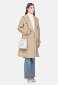 Cappotto in lana beige con colletto a scialle, abbinato a jeans flare azzurri chiari, mentre tiene una borsa a secchiello in pelle bianca con hardware dorato.