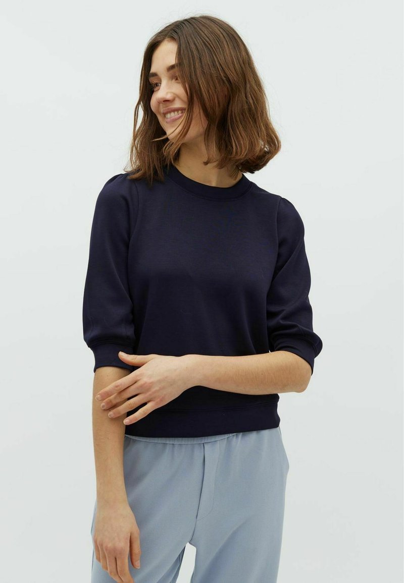Maglione blu navy con collo rotondo e maniche a sbuffo, realizzato in materiale morbido. Abbinato a pantaloni azzurri chiari, larghi.
