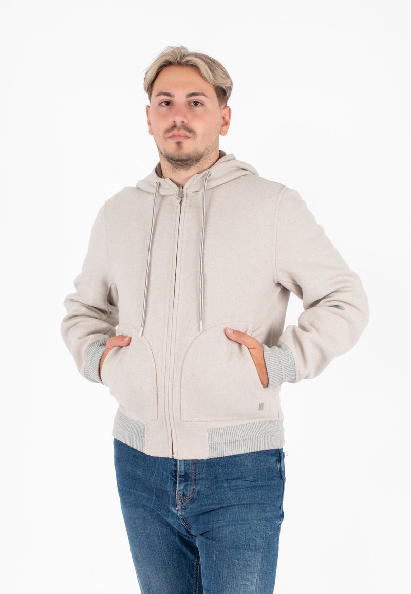 Felpa con zip di un beige chiaro, dotata di cappuccio, tasche frontali e polsini a coste grigi. Realizzata in un tessuto morbido, presenta una superficie liscia e un design minimalista.