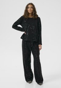 Completo top e pantaloni neri con paillettes, caratterizzato da una vestibilità ampia e dettaglio peplum. Superficie testurizzata con accenti lucidi; il modello indossa scarpe in contrasto.