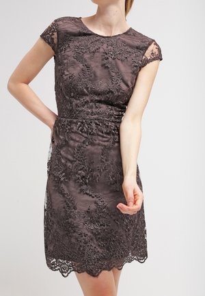 Vrouw die een donker kanten jurk draagt, kort mouwtje en tot aan de knie, met bloemrijke borduursels en een gegolfde zoom, staand tegen een effen achtergrond.