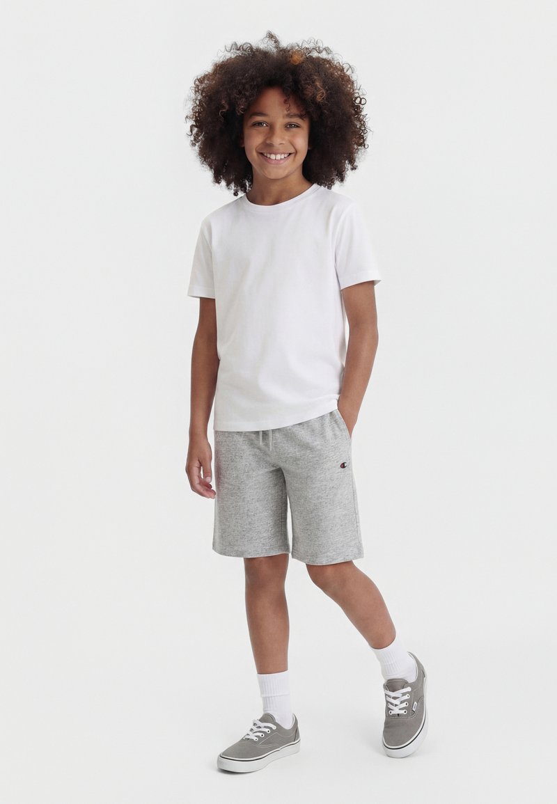 Enfant aux cheveux bouclés souriant, portant un t-shirt blanc, un short gris, des chaussettes blanches et des baskets grises, debout devant un fond uni.