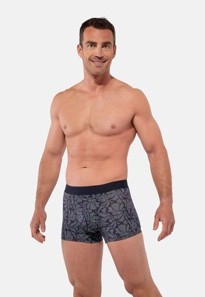 Muskulöser Mann lächelt, trägt gemusterte dunkle Boxershorts mit einem schwarzen Bund, steht vor einem schlichten hellen Hintergrund.