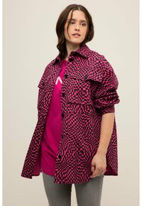 Chaqueta de camisa a cuadros rosa y negra con mangas largas, dos bolsillos en el pecho y cierres de botón. Llevada sobre una camisa rosa a juego. Jeans grises.