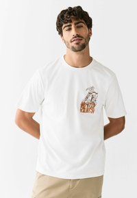 Valge puuvillane T-särk, millel on graafiline trükk ratsanikust hobusel ja "OUTDOOR RANCH" oranžides toonides vasakul rinnal. Lühikesed varrukad.