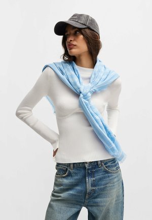 Heller blauer Schal mit Logo-Muster, drapiert über einem weißen, gerippten Langarmoberteil, kombiniert mit blauen Jeans und einer dunklen Mütze.