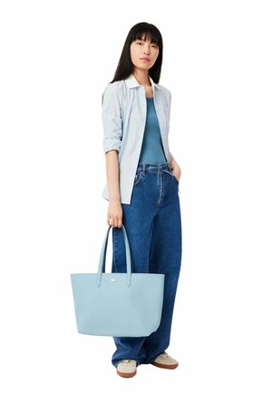 Sac fourre-tout bleu clair avec une finition texturée et des poignées doubles, agrémenté d'un petit logo. Le modèle porte un débardeur bleu et une chemise à rayures ajustée.