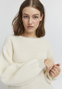 Pull en tricot crème clair avec texture côtelée, col rond et manches longues, offrant une coupe décontractée et une finition douce.