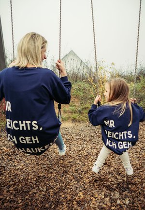 Zwei Personen auf Schaukeln, die passende dunkelblaue Sweatshirts mit weißem Text tragen, umgeben von Holzschnitzeln in einem Freien.