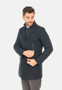 Cappotto nero su misura con colletto a risvolto, bottoni frontali e tasche laterali con zip. Realizzato in tessuto liscio e strutturato, design di lunghezza media.