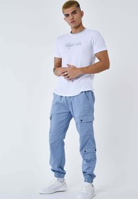 Giovane con capelli corti tinti, che indossa una maglietta bianca "Project x Paris", pantaloni cargo azzurri chiari e sneakers bianche, in piedi con le mani unite.