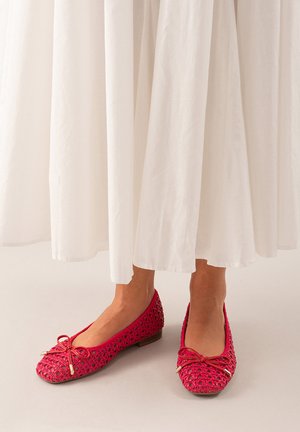 Zapatos de ballet de color rosa brillante, hechos de un material entrelazado, con un gran lazo en la parte delantera y una suela plana, llevados con una falda blanca.