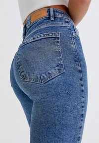 Jeans de mezclilla azul de cintura alta con un acabado desgastado, que presentan un bolsillo trasero y detalles de parche de cuero en la cintura.