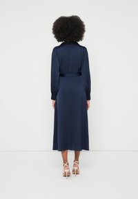 Marineblå satengkjole med innsvingt midje, lange ermer og krage. Kjolen har et flytende skjørt og er kombinert med lette, remmede hæler.