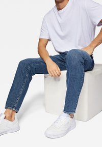 Hombre sentado en un cubo blanco, vistiendo zapatillas blancas, jeans azules y una camiseta blanca de manga corta sobre un fondo liso.
