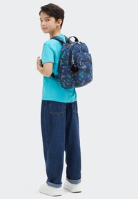 Modrý látkový batoh s vzorem vesele se usmívajících dinosaurů, přední zipová kapsa, nastavitelné popruhy a měkký černý přívěsek připevněný na zipu.