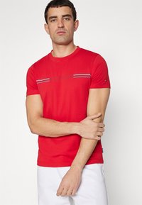 T-shirt di cotone rossa a maniche corte, con dettaglio a righe orizzontali in blu e bianco sul petto e un piccolo logo sulla manica.