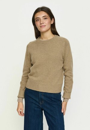 Femme aux longs cheveux bruns portant un pull beige et un jean bleu, debout devant un fond clair uni, faisant face et souriant légèrement.