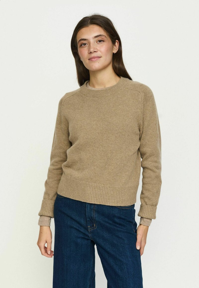 Femme aux longs cheveux bruns portant un pull beige et un jean bleu, debout devant un fond clair uni, faisant face et souriant légèrement.