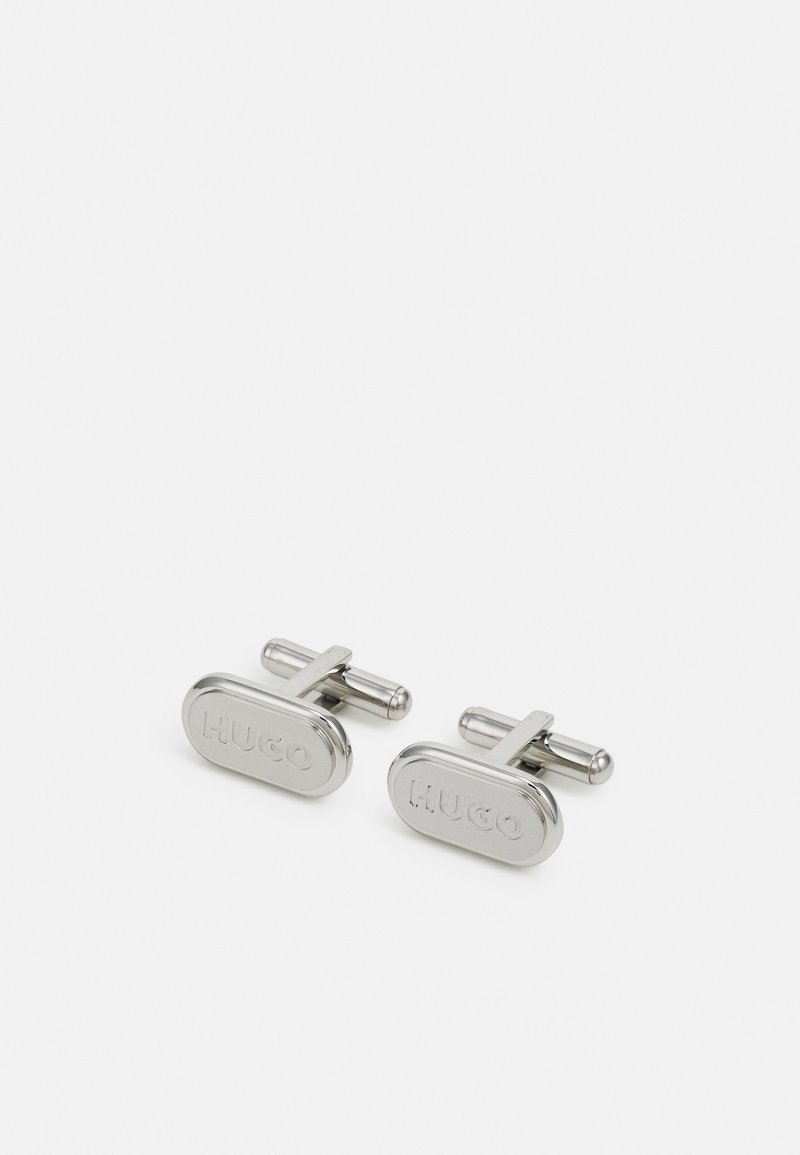 Chaussettes en argent avec une forme ovale présentant un logo "HUGO" en relief. Fabriquées en métal poli, elles ont un design minimaliste et une texture lisse.