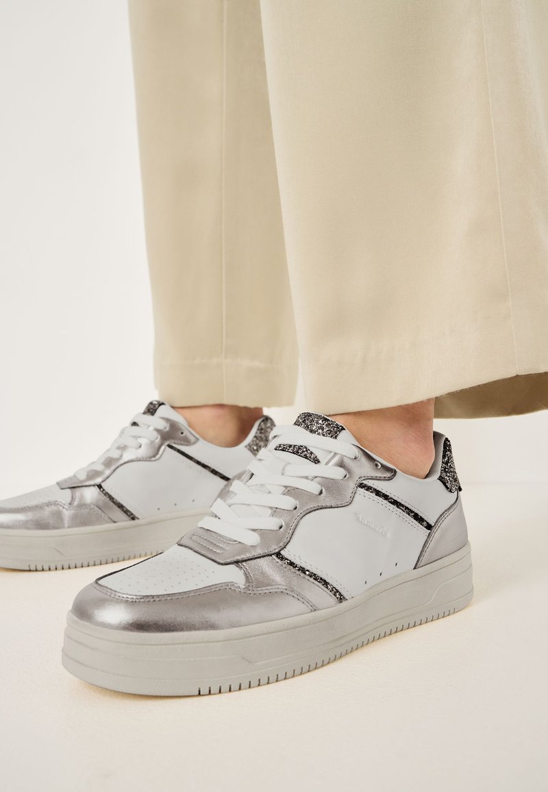 Zilveren en witte low-top sneakers met gestructureerde zilveren accenten, een zwarte glinsterende hiel detail, en witte veters op een lichte rubberen zool.