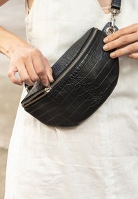Borsa in pelle nera con texture a coccodrillo, forma ovale, doppia cerniera e dettagli hardware argentati. Da portare a mano su uno sfondo chiaro.