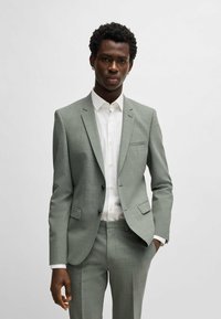 Costume gris ajusté avec une finition texturée, comprenant une veste à un bouton, des revers en côtelettes et un pantalon plat associé à une chemise blanche.