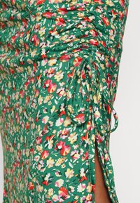 Green floral fabric with small red, white, and yellow flowers, featuring gathered ruched detailing and tied drawstrings on a side slit.